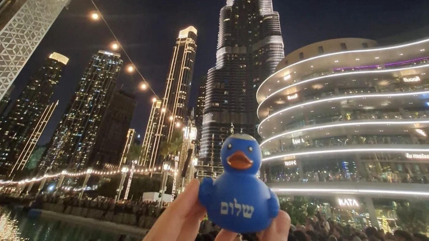 A hand holds up a blue Kosher Duck Revolution rubber duck with שלום on its chest against a glittering night-time city skyline